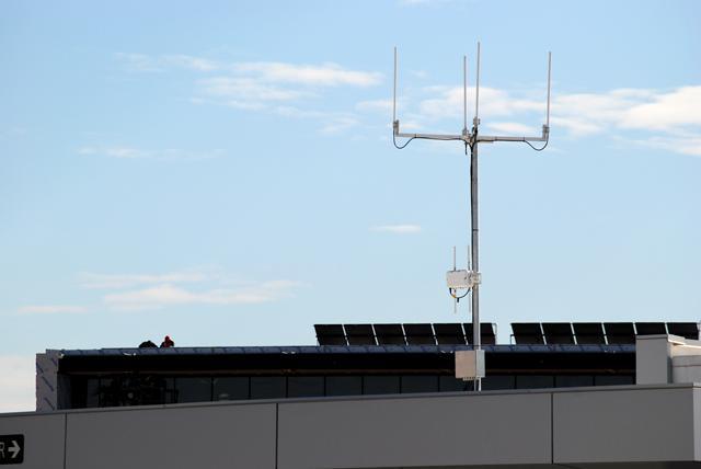 This CentMesh node is located atop the Centennial Parking Deck, across the road from the Hunt Library, where construction workers work on the roof. Each node consists of two parts. The top antenna array, connected to a computer on the ground, is part of the CentMesh network, transmitting and receiving signals. The other small box is a commercial Cisco router, which will be used to create a network that the open source CentMesh network can be compared to. Photo by Michael Pratt