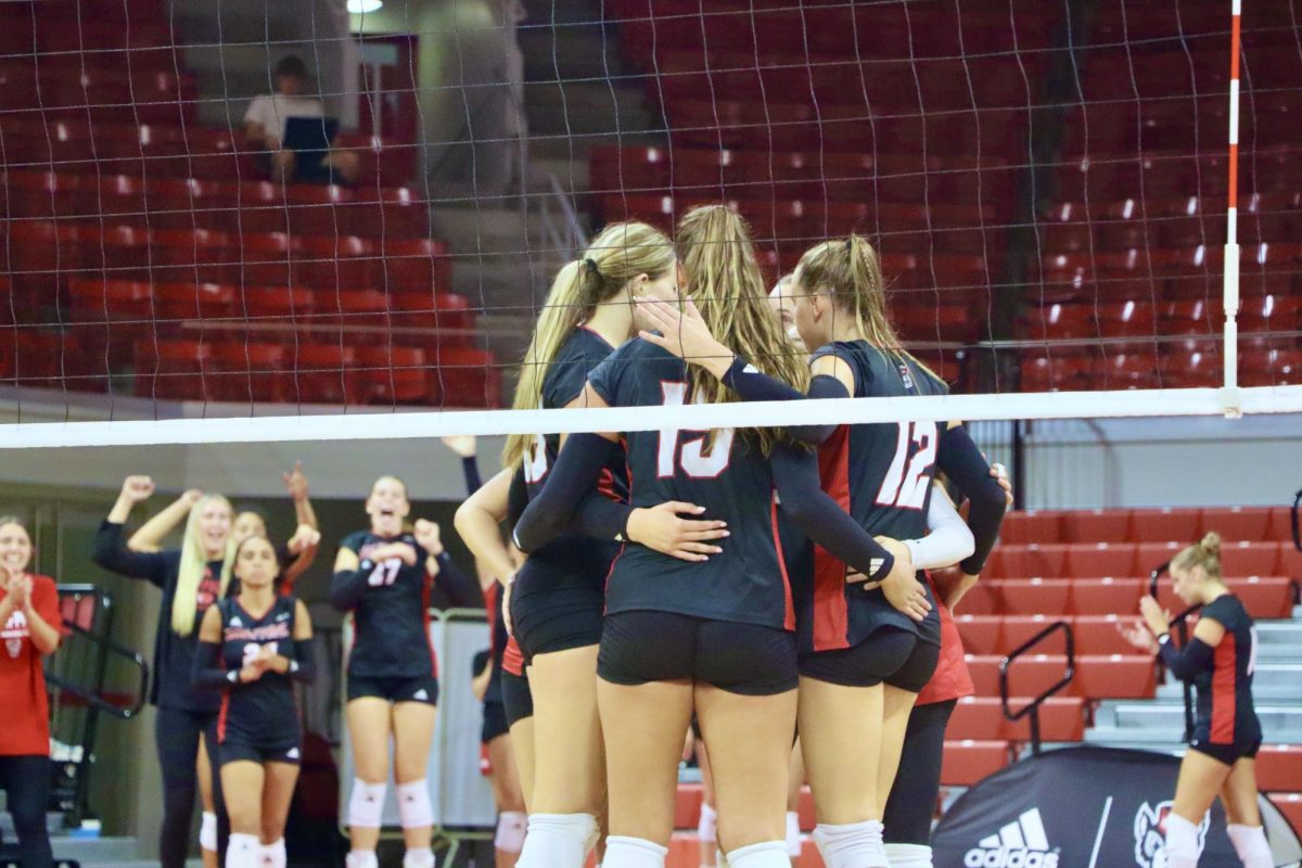 NC State players huddle on the court while their teammates cheer in the background during the game against Akron at Reynolds Coliseum on Friday, Aug. 29, 2025. The game was the first time the Wolfpack and the Zips have faced off in women’s volleyball since 2008. NC State beat Akron 3-2.